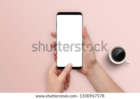 Female hands holding modern smartphone with empty screen on pink desk background with coffee, Mockup