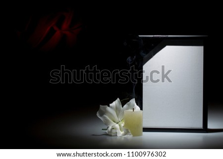 Blank black mourning frame, with smoky candle and white lily flower on dark background with red decoration