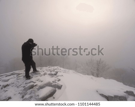 Photographer takes picture of freeze snowy mountains rocks. Extreme misty weather. Stony rock peak increased from foggy valley.