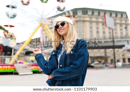 Incredible happy young woman with lovely smile dressed denim jacket and white blouse, black sunglasses posing in the city on background of amusement park