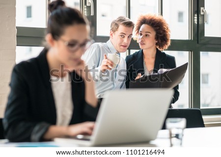 Two young malicious employees gossiping about their hard-working colleague in the office Royalty-Free Stock Photo #1101216494