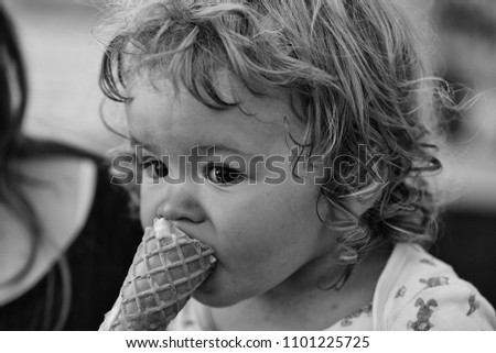 Portrait of small hungry funny baby boy with wet blonde curly hair eating cold tasty ice cream in wafer from hands of mother outdoor, horizontal picture