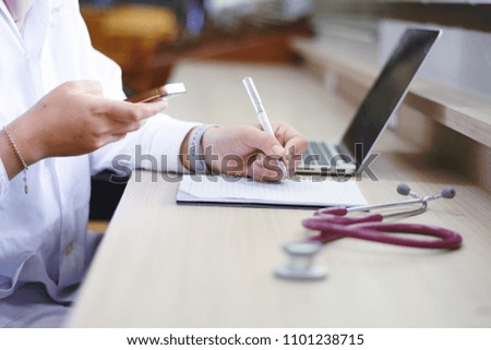 Asian woman doctor writing in a note and using smart phone