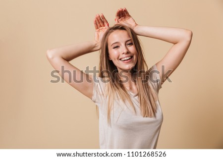 Image of lovely blonde girl 20s in casual t-shirt smiling and showing rabbit ears at her head isolated over beige background
