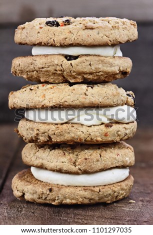 Stack of three Carrot cake raisin cookies sandwiches stuffed with cream cheese icing against a rustic wood background.