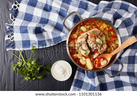 delicious rich pork vegetable soup with white beans, green peas, red bell pepper, herbs, species in a metal casserole with wooden spoon, on a black wooden table, view from above, close-up