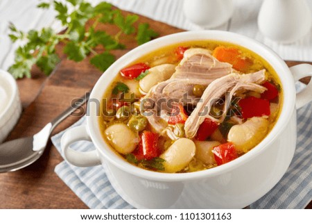 summer pork vegetable soup with white beans, green peas, red bell pepper, herbs, species in a white bowl with silver spoon and sour cream on a wooden board, view from above, close-up