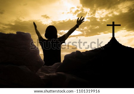 Happy woman standing with hands up on rocks with silhouette wood cross on sunset background, success and hope concept