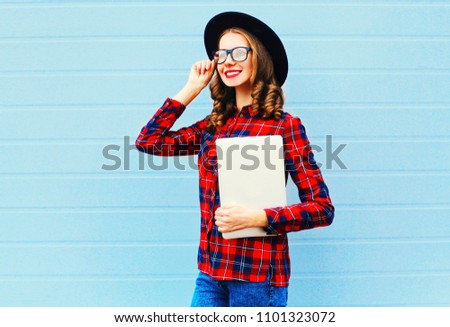 Stylish woman holding laptop computer or tablet pc in city, wearing black hat, red checkered shirt over blue background