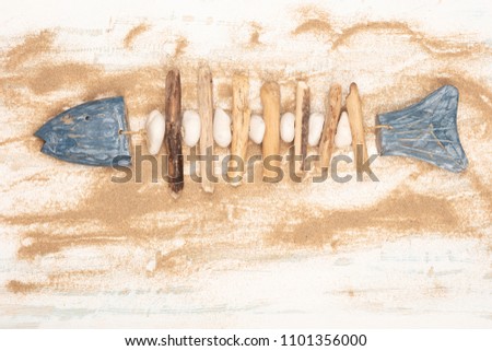 Decorative hand-crafted fish formed of stones and wood on scattered beach sand over a white and blue wooden   background for nautical or marine concepts