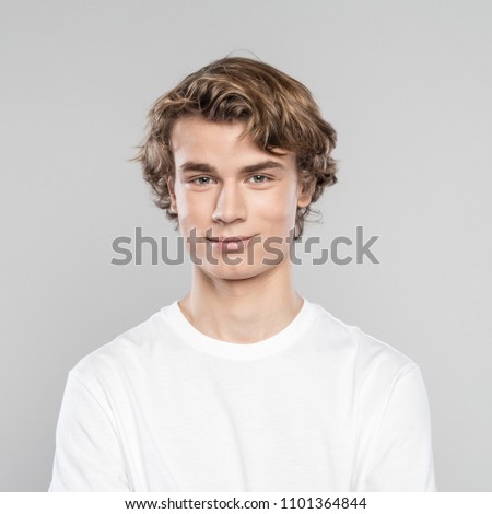Studio shot of cheerful teenage boy wearing white t-shirt, standing against grey background, smiling at camera.