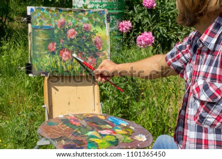 A bearded male artist paints painting in nature. Plein air drawing a bush of beautiful peony flowers rural landscape