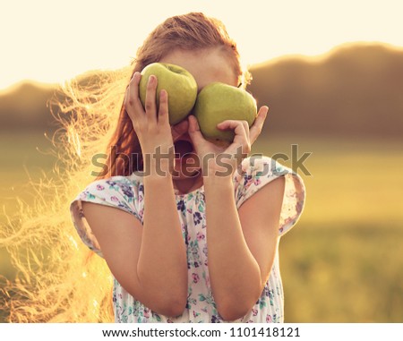 Fun surprising beautiful kid girl with long hair playing with green apples on summer bright background. Closeup toned color portrait
