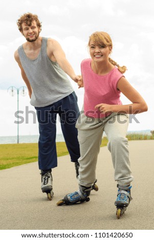 Active holidays, exercises, relationship concept. Young woman dressed in sports clothes putting her boyfriend up to do rollerblading while holding his hand on promenade