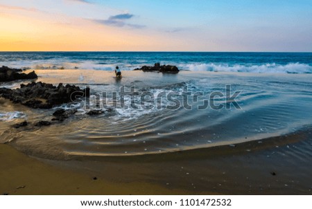 Wide host of a beach in the Dominican Republic with a ten year old boy playing in the shallow tidal zone. Ripples of water flow inland created by the rocks. The sun is setting.