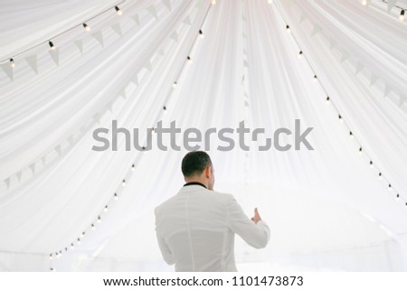 Young man in white jacket standing on the background of white tent with lanterns garland. Show begins. Back view