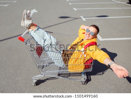 fashion guy in sunglasses and a yellow jacket sits in a cart from food in the supermarket parking