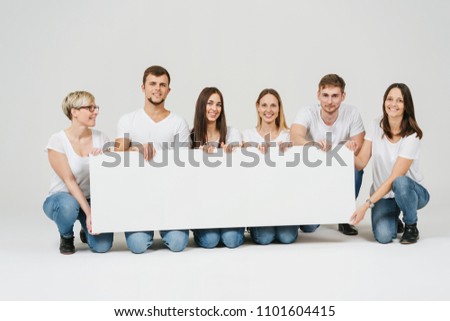 Happy friendly group of friends in jeans and t-shirts kneeling with a blank white panorama banner in front of them over a white studio background