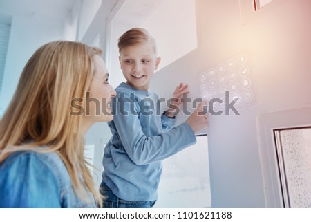 Curious child. Cheerful kind boy touching a modern device on the wall of his smart house and looking attentively at his mother