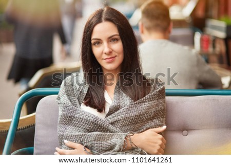Pretty brunette female with healthy skin and pleasant apperance, sits in outdoor restaurant, wrapped in coverlet, looks directly and calm at camera, enjoys good atmopshere in cozy sidewalk cafe