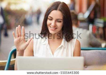 Positive Caucasian female makes video call via laptop computer, waves with hand as sees relatives from abroad, share news, pose against outdoor coffee shop background, uses free interent connection