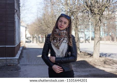 Young cute girl with blond hair in the city at the road looks at the camera. A girl in a black leather jacket and with glasses on her head