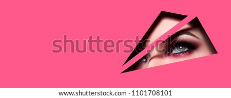 The face of a young beautiful girl with a bright make-up and with plump red lips peeks into a hole in pink paper.Fashion, beauty, make-up, cosmetics, beauty salon, style, personal care, geometry.