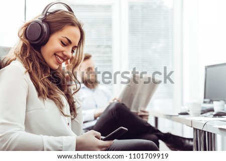 Businesswoman with headphones sitting at desk during the break and using mobile phone while man is reading newspaper Royalty-Free Stock Photo #1101709919