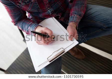 Man writes in a notebook on his knee while sitting on steps. Close up picture taken from high angle of male hand writing on blank white paper on a clipboard sitting down.