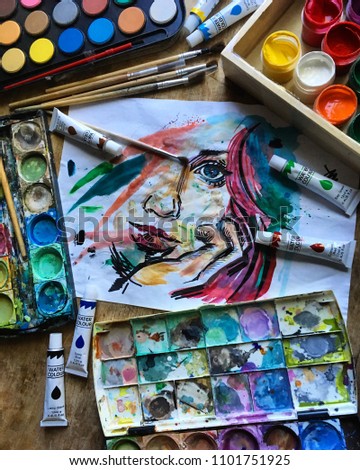The girl's face is painted in watercolor. Wooden table, gouache, watercolor, notebook, brushes, oil paint, wooden box. The working process. Close up.