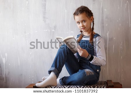 Young cute emotional teenager girl with braids in overalls portrait indoor. Sitting on chest and reading book.