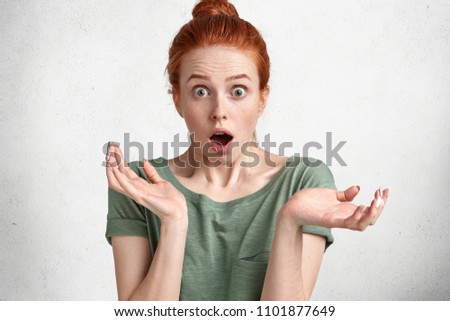 Puzzled emotive red haired Caucasian female shrugs shoulders with frustration, has shocked expression, can`t believe her eyes, wears casual t shirt, isolated on concrete wall. Clueless ginger woman