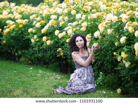 Young brunette Caucasian woman with curly hair sitting on green grass near yellow roses Bush in a garden, looking straight to the camera