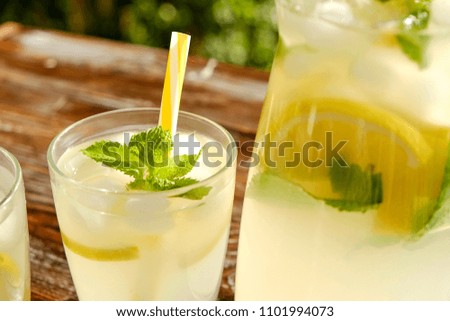 Lemonade pitcher and two glasses of cold refreshing citrus beverage with ice, lemon slices, mint leaves & straws on brown grunged wooden table, country side foliage background. Close up, copy space.