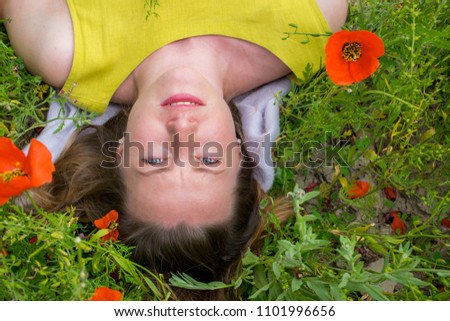 European caucasian happy young woman, blonde with open shoulders in a yellow dress with red lips and nice smile lies in the field of the blossoming red poppies..