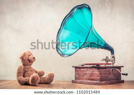 Retro Teddy Bear toy sitting near antique classic old gramophone turntable player on oak wooden floor front concrete wall background. Listening to music concept. Vintage instagram style filtered photo