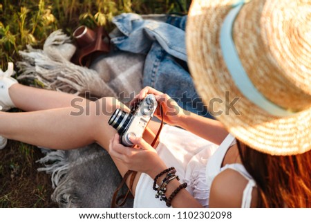 Cropped image of european woman wearing straw hat holding retro camera while sitting on grass in summer park