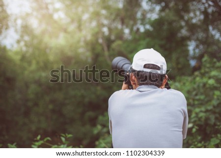 A man, standing in front of a forest covered with green trees, holds up a camera with a long lens that shines out to capture his memories of the day.