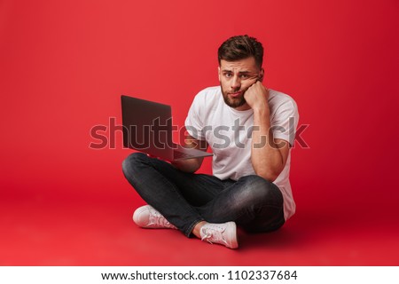 Photo of displeased bored man in t-shirt and jeans sitting on floor and propping his head with hand while using laptop isolated over red background