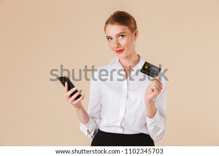 Photo of young business woman standing isolated over beige wall backgound looking camera using mobile phone holding credit card.