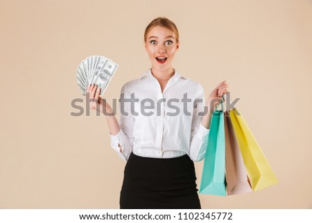 Picture of excited young business woman standing isolated over beige wall backgound looking camera holding shopping bags and money.