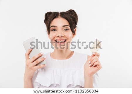 Image closeup of happy woman 20s with double buns hairstyle and dental braces looking at you while holding smartphone and credit card in both hands isolated over white wall