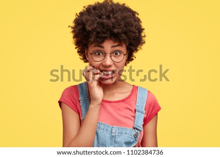 Frustrated beautiful African American female feels anxious and surprised, bites finger nails in puzzlement, looks nervously at camera, worries before presenting project work in front of audience Royalty-Free Stock Photo #1102384736