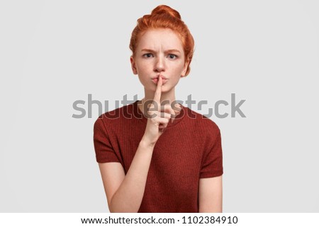 Angry beautiful red haired female with sullen expression, demands silence on exam, asks not to cheat, frowns face, shows secret sign, stands against white background. Don`t make too much noise! Royalty-Free Stock Photo #1102384910