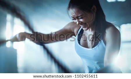 Athletic Female in a Gym Exercises with Battle Ropes During Her Cross Fitness Workout/ High-Intensity Interval Training. She's Muscular and Sweaty, Gym is in Industrial Building. Royalty-Free Stock Photo #1102455611