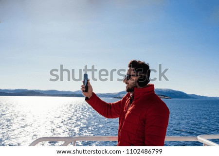 Side view of handsome caucasian man in red winter jacket making photos on the smartphone camera. Beautiful sea view from the boat. Norway