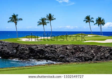 Sunny day on a tropical golf course fairway with the putting green in the distance surrounded by palm trees and sand traps, lava rock, blue pacific ocean, and blue sky and white clouds in background Royalty-Free Stock Photo #1102549727