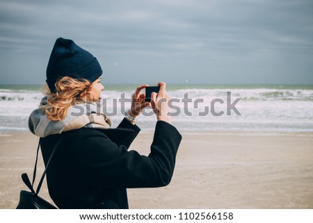 Happy young woman traveler making picture of seashore on her smart phone on cold season. Stylish hipster female smiling having fun on ocean beach at sunny day.