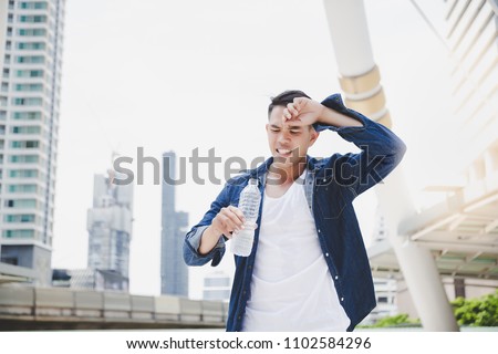 Attractive handsome asian man gets thirsty because of hot weather in the summer season. Cool guy feels exhausting and holds the bottle of cold water. He wears denim jacket. copy space, city background Royalty-Free Stock Photo #1102584296