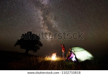 Night camping near silhouette of big tree on grassy valley. Tourist man standing near burning campfire, pointing at beautiful starry sky and Milky way to young woman sitting in glowing tent entrance
